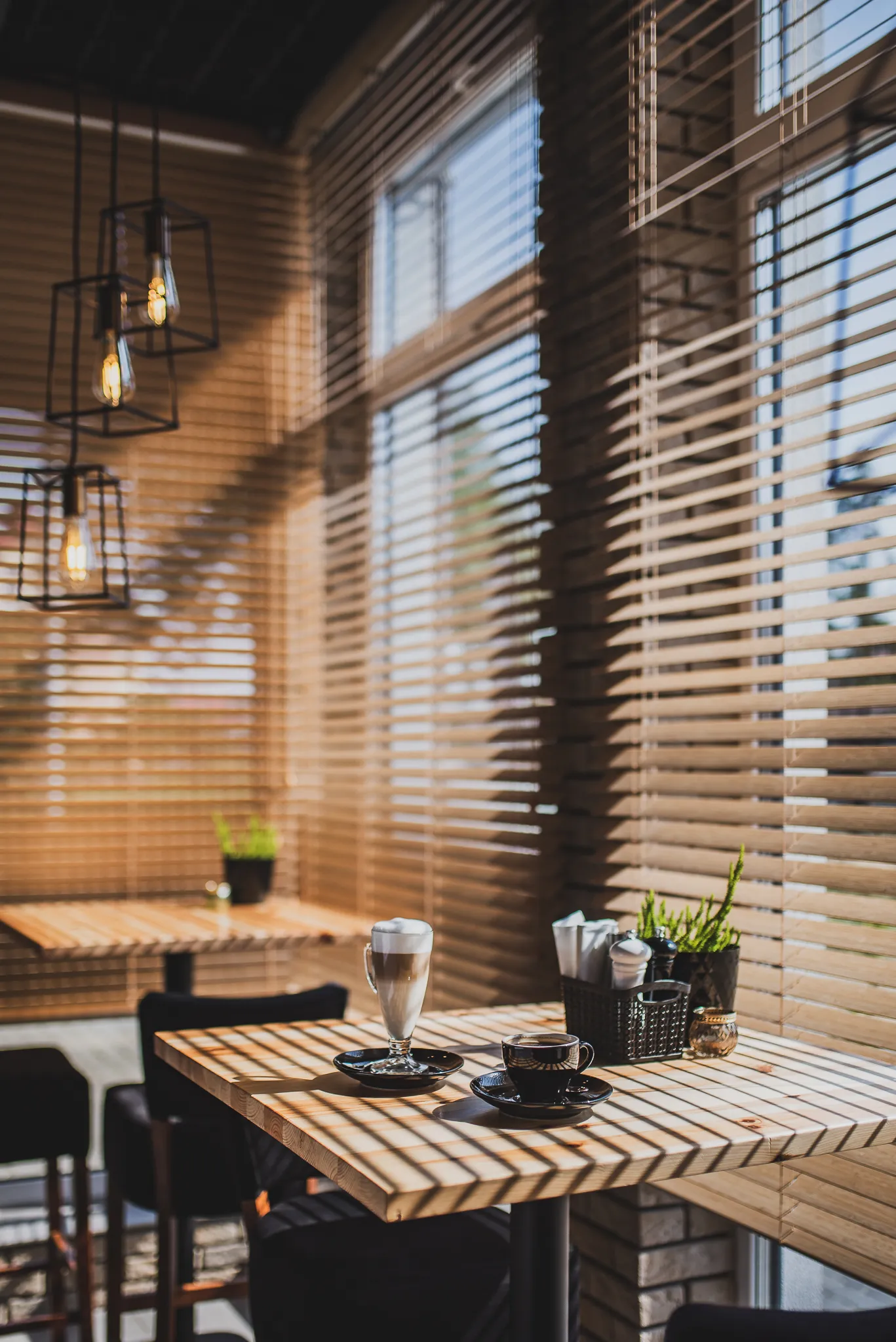 Stolik z filiżanką i szklanką kawy na tle okien z żaluzjami i ceglanej ściany.