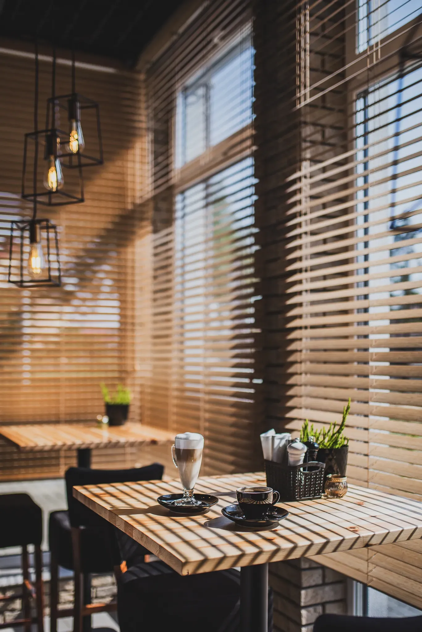 Stolik z filiżanką i szklanką kawy na tle okien z żaluzjami i ceglanej ściany.