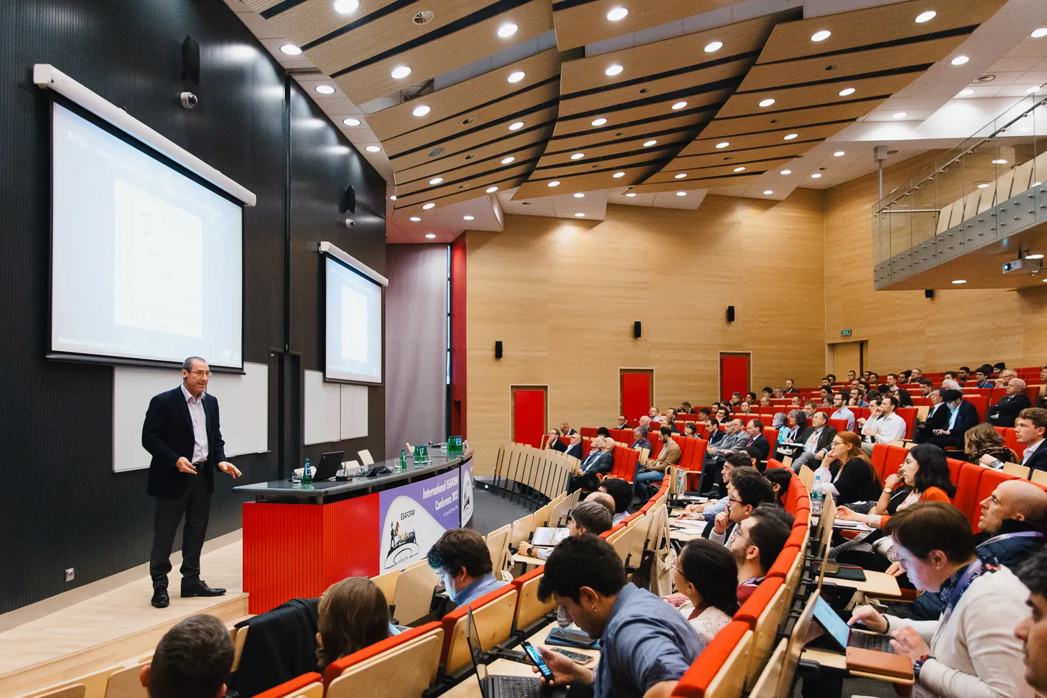 Mężczyzna w garniturze stoi z przodu sali wykładowej, zwracając się do dużej publiczności siedzącej w rzędach czerwonych krzeseł.