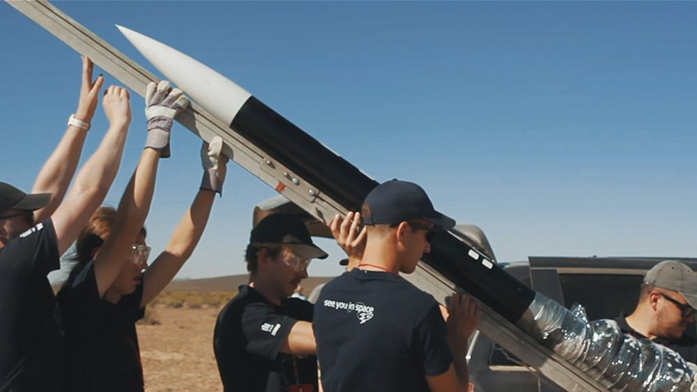 Grupa ludzi pracuje razem, aby podnieść dużą rakietę w powietrze, z widocznym biało-czarnym korpusem i spiczastym czubkiem rakiety.