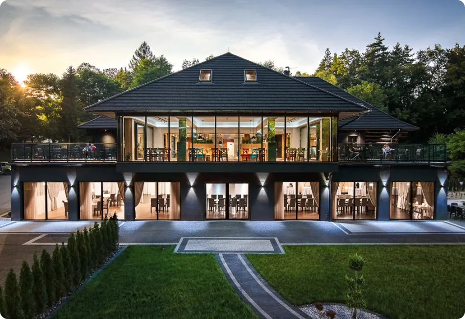 Duży, nowoczesny dom z przestronnym podwórkiem i balkonem na drugim piętrze, z dużą przeszkloną ścianą, z której roztacza się widok na wnętrze. Dom położony jest w zalesionej okolicy, z drzewami widocznymi w tle.